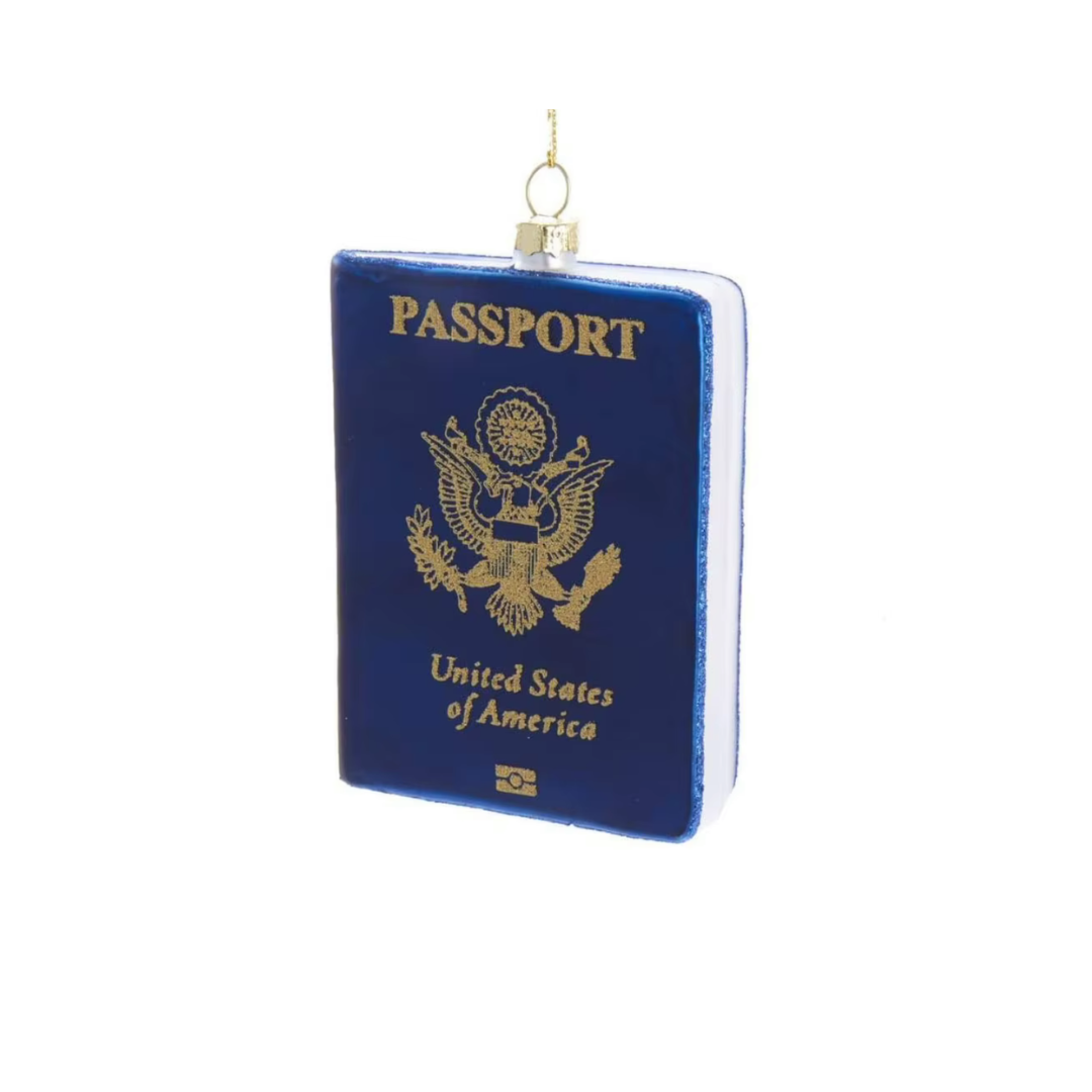A novelty Christmas ornament shaped like a United States passport, featuring a dark blue body with gold foil-style lettering and emblem, hanging from a small gold loop on a white background.