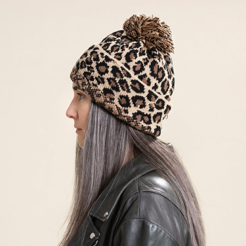 A woman in profile wearing a leopard print knit beanie with a brown pom-pom, paired with a black leather jacket, against a solid light beige background.