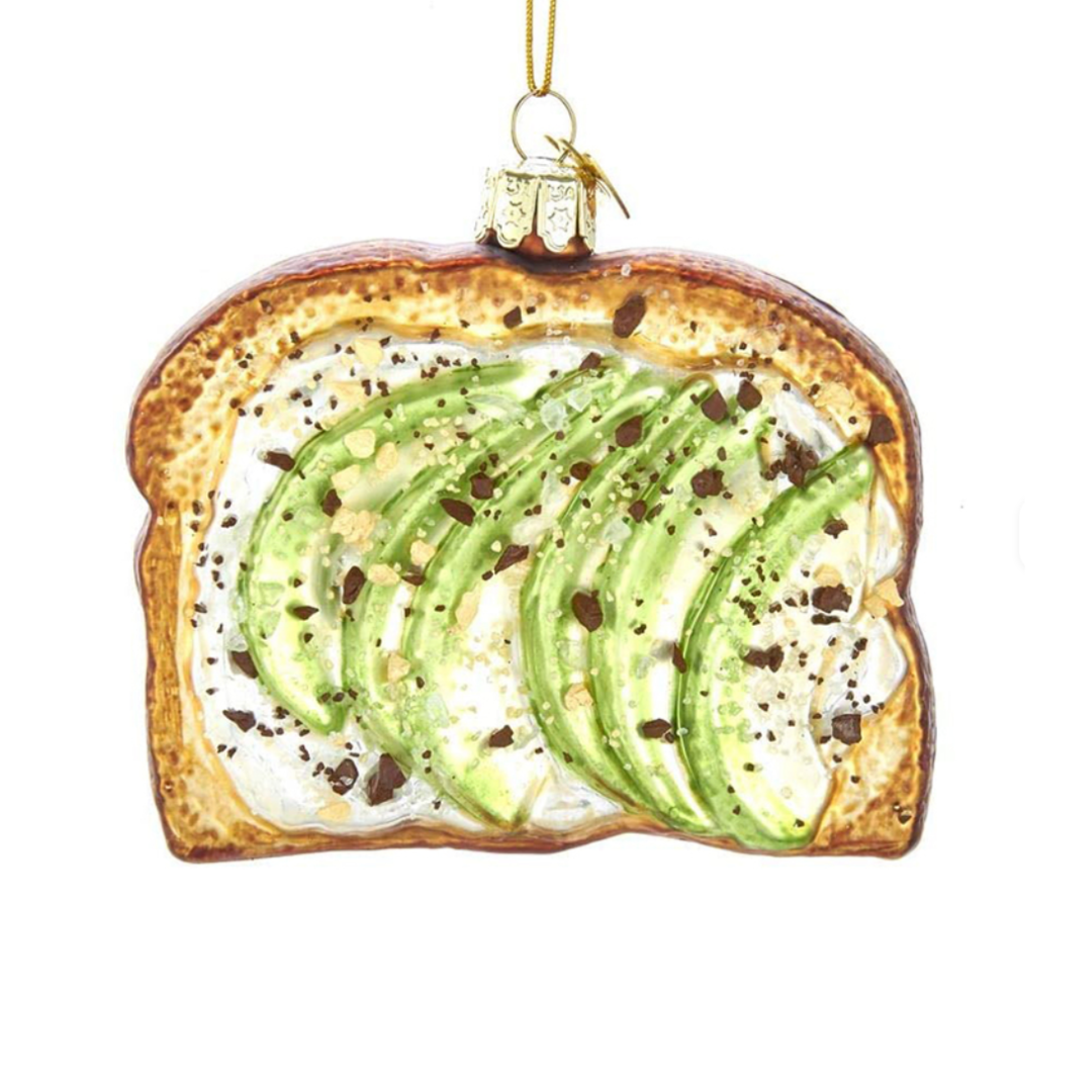 A toast-shaped ornament featuring avocado slices, black pepper flakes, and a golden-brown toasted crust, hanging from a gold string on a white background.