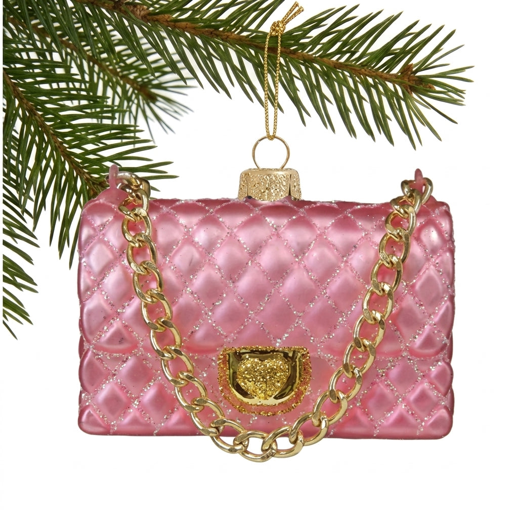 A pink quilted handbag Christmas ornament with gold-tone chain and clasp details, hanging from a pine branch against a white background.